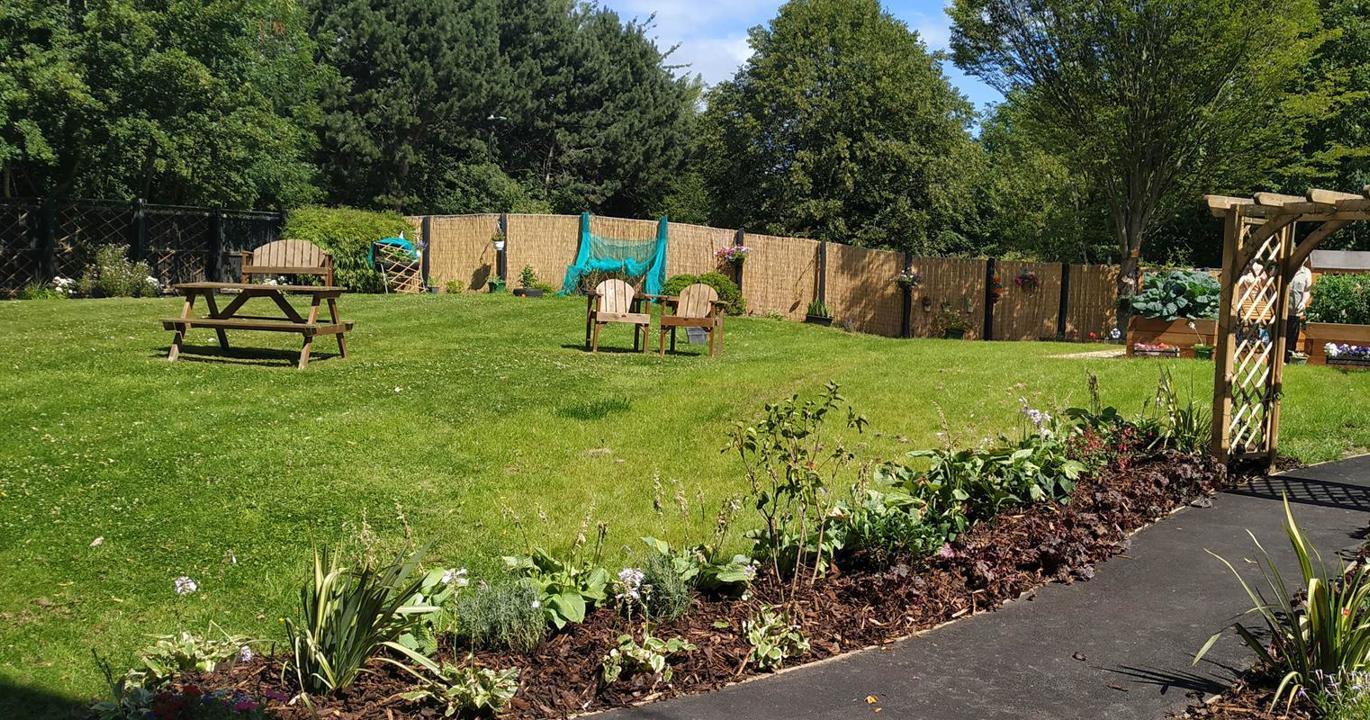 A lovely green garden with wooden furniture in the background, a curved tarmac walkway bordered by flower beds in the foreground