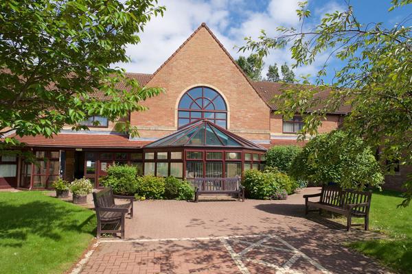 The entrance to Swan Court, a welcoming building with two wooden benches and a wide accessible walkway borderd by lush green lawns