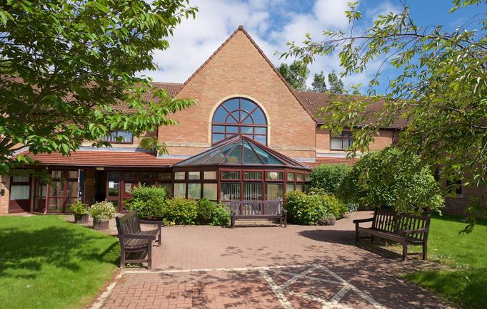 The entrance to Swan Court, a welcoming building with two wooden benches and a wide accessible walkway borderd by lush green lawns