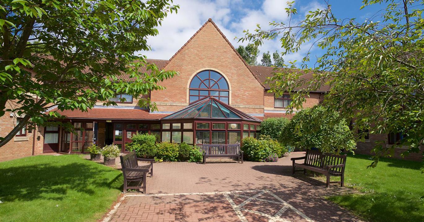 The entrance to Swan Court, a welcoming building with two wooden benches and a wide accessible walkway borderd by lush green lawns