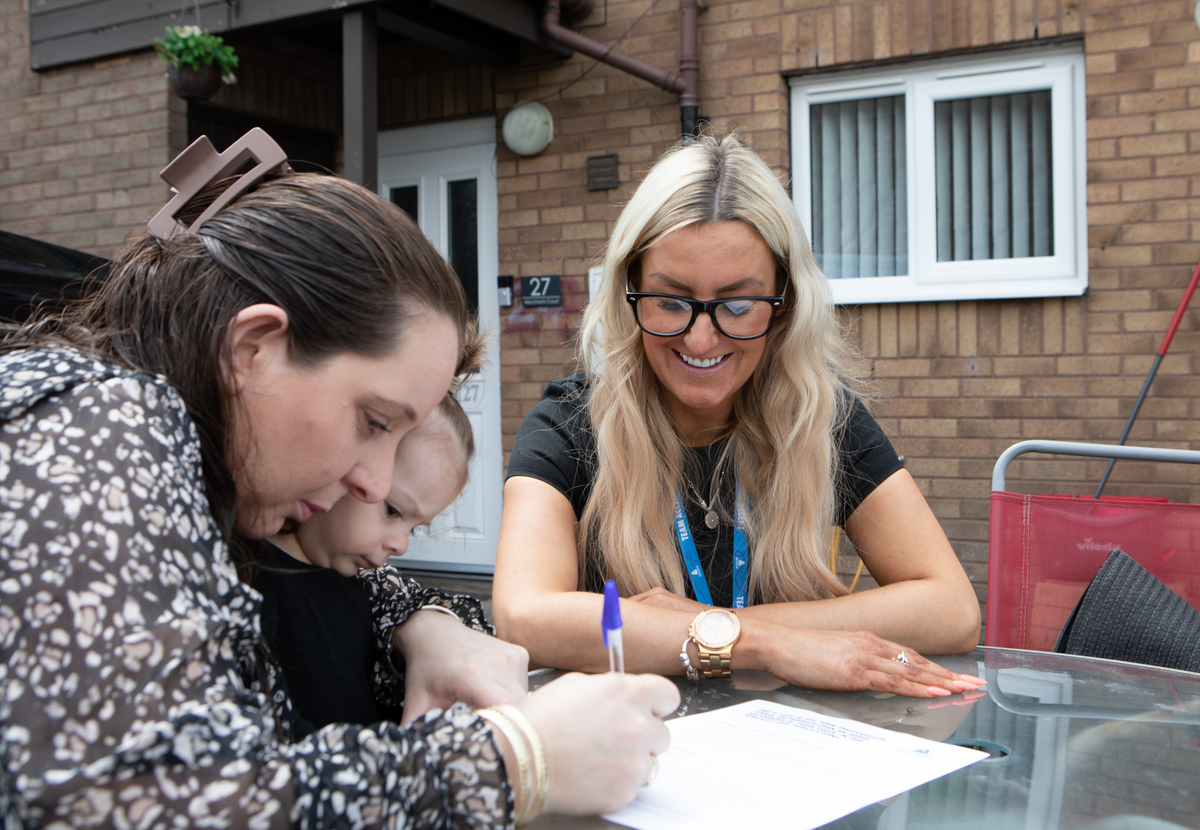 Two women sit at an outdoor table while one signs a piece of paper.