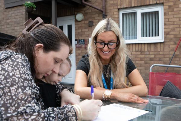 Two women sit at an outdoor table while one signs a piece of paper.