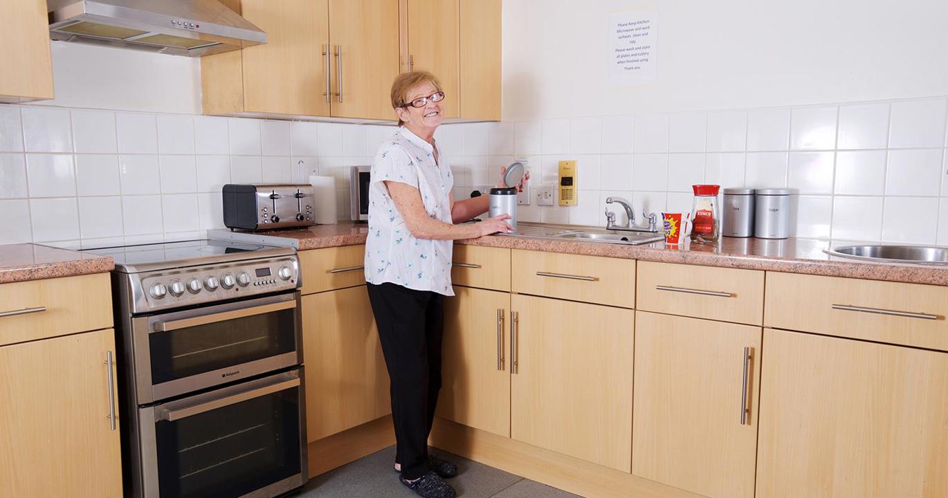 A smiling woman opens a coffee jar in a kitchen with an oven, sink, toaster and other amenities