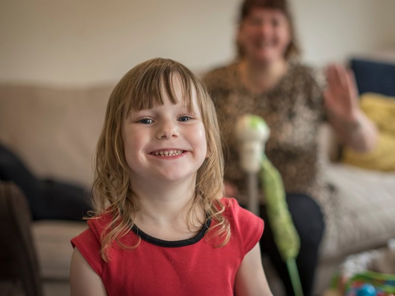 A young child in a red teeshirt smiling broadly from the inside of her home