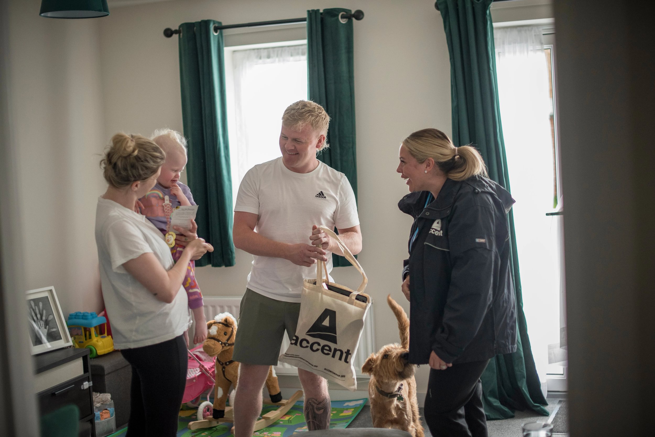 Three people crowd around a baby in a living room – everyone is smiling, and there's a dog wagging its tail 