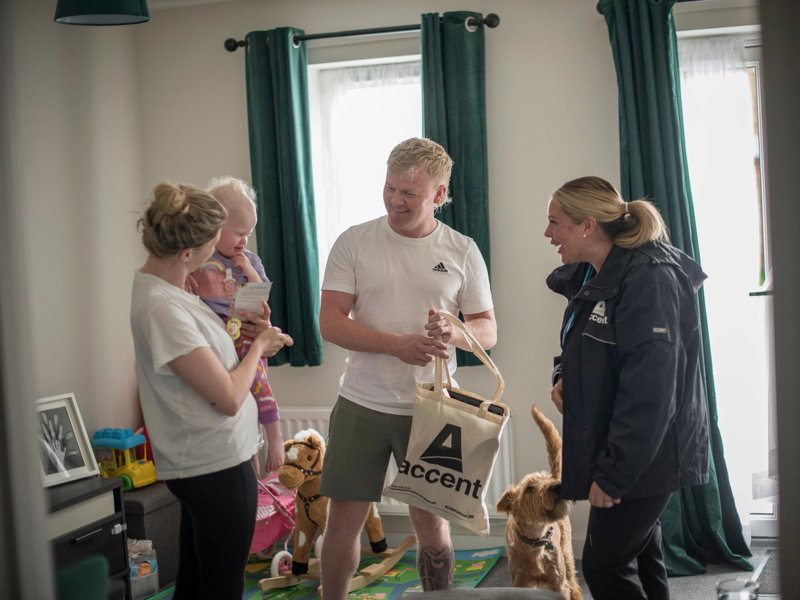 Three people crowd around a baby in a living room – everyone is smiling, and there's a dog wagging its tail 