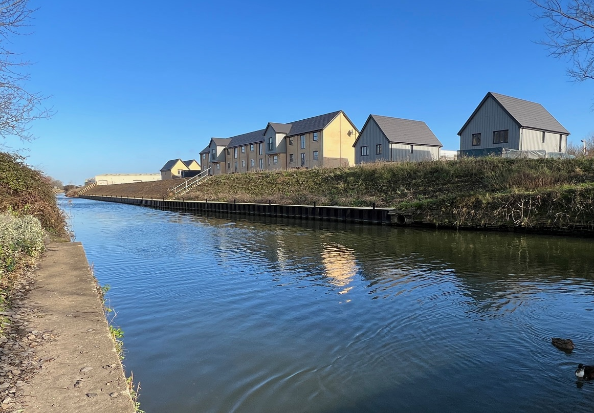 A canal on a sunny day, with a number of newly built homes on the far bank