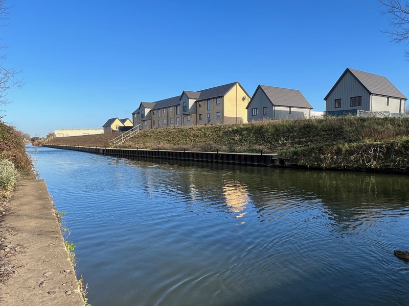 A canal on a sunny day, with a number of newly built homes on the far bank