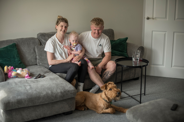 A family of three sitting on a grey sofa with a shaggy brown dog at their feet