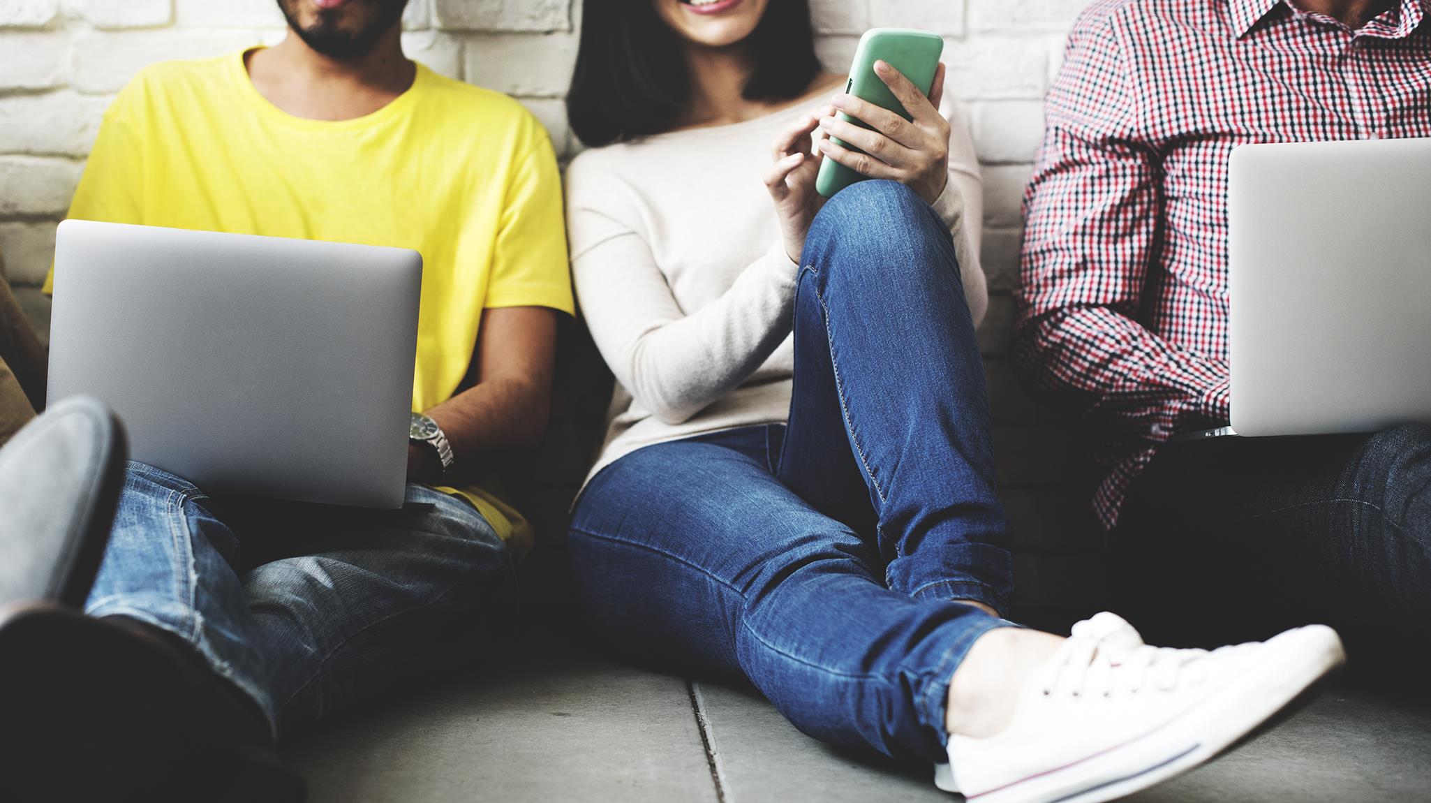 A group of people wearing casual clothes sitting on the floor using electronic devices