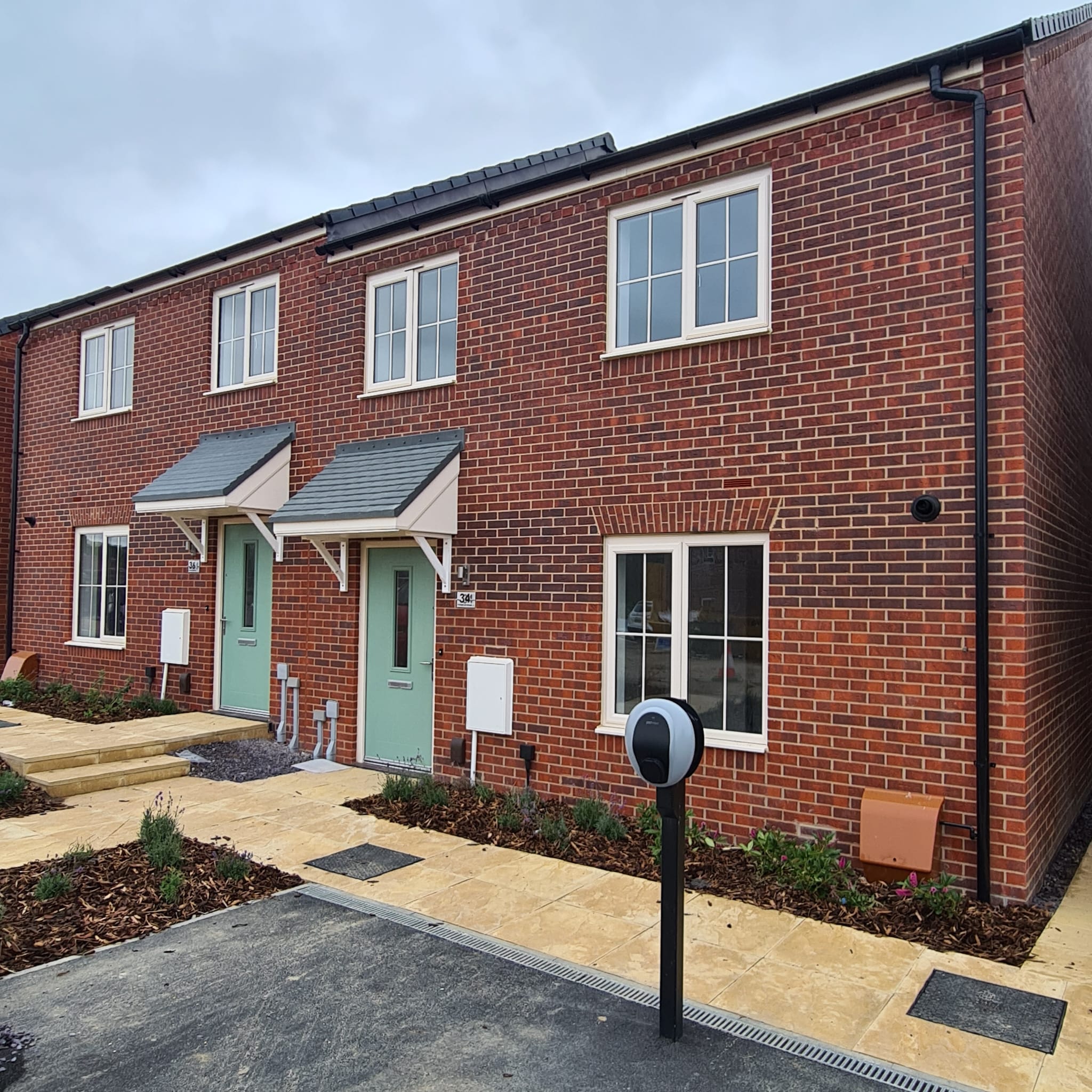 A two-storey red brick housing development with a sage-coloured front door and newly planted flower beds