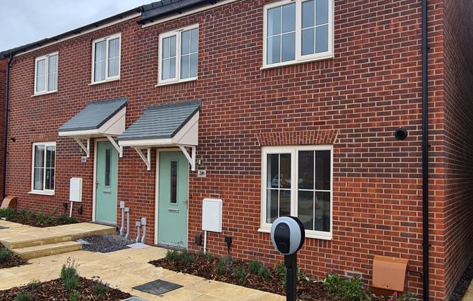 A two-storey red brick housing development with a sage-coloured front door and newly planted flower beds