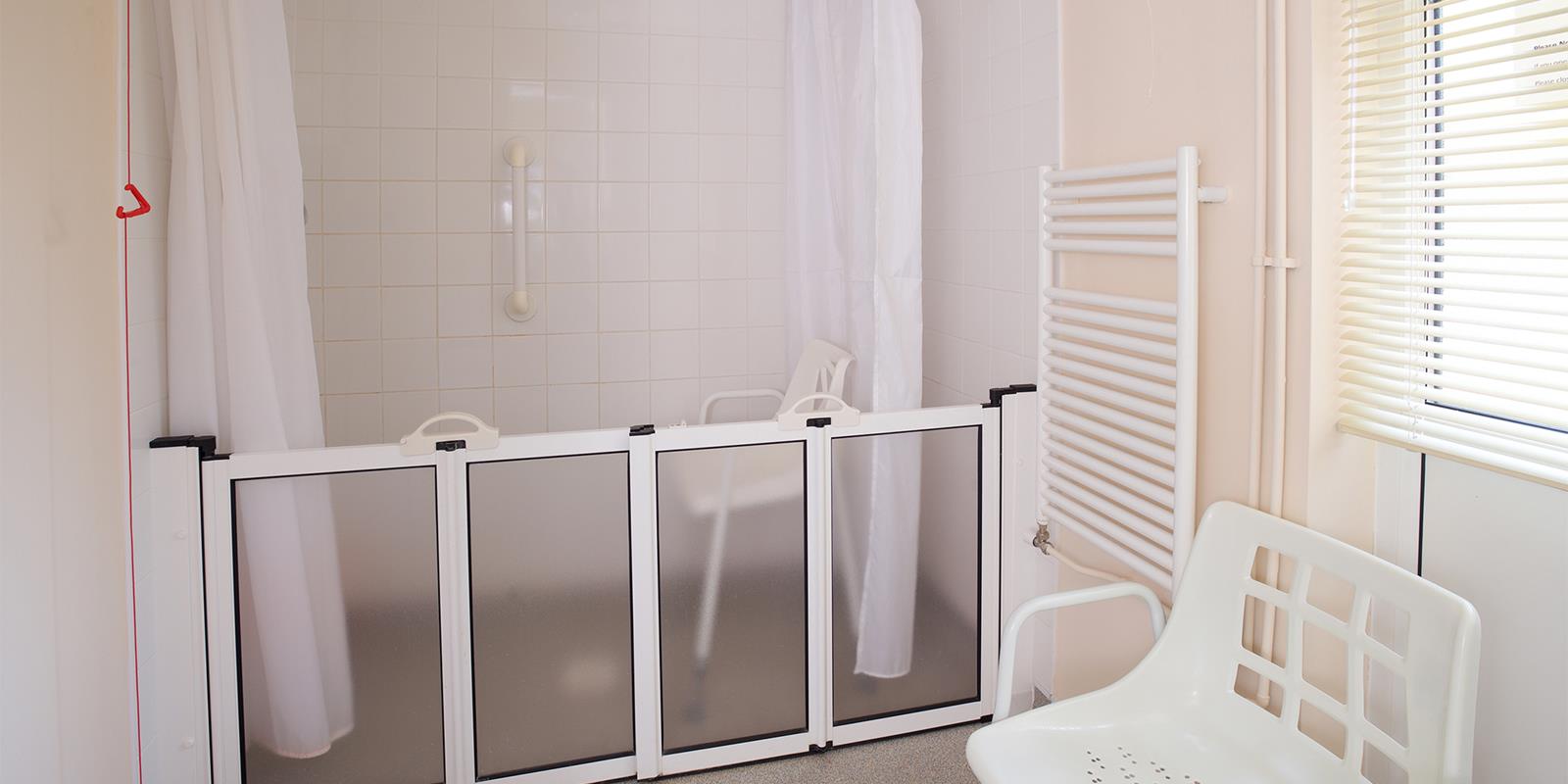 A shower-room with a walk-in shower, featuring a retractable plastic gate and a white plastic chair in the foreground