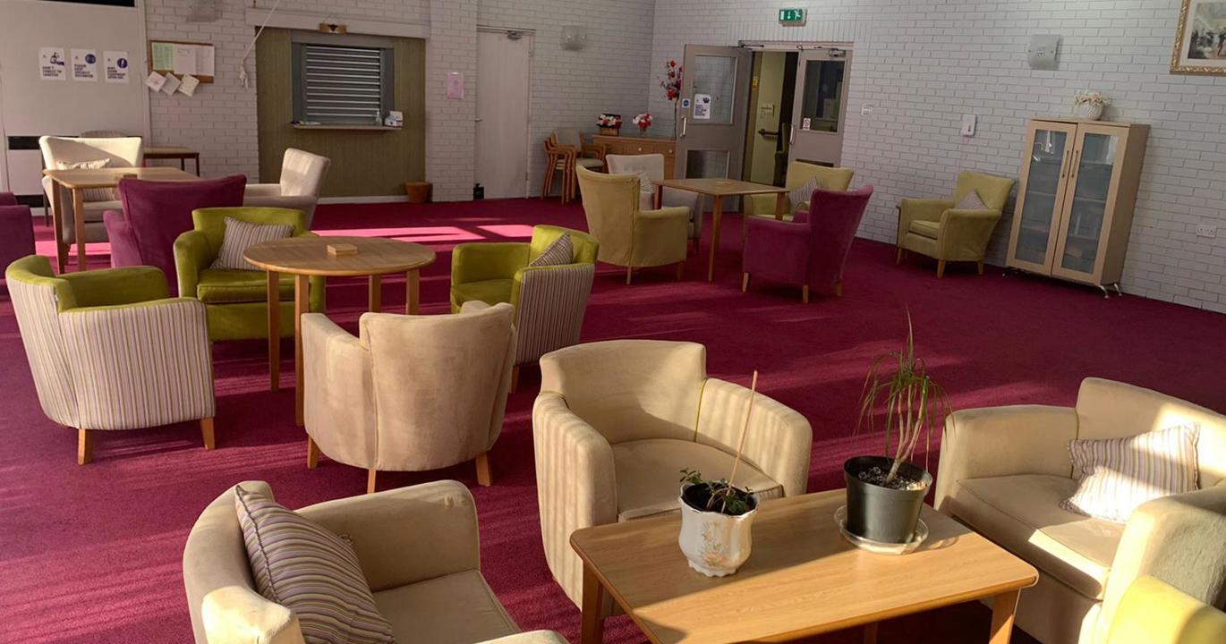 A sun-lit recreation room with a red carpet, white walls and a number of comfortable chairs surrounding coffee tables